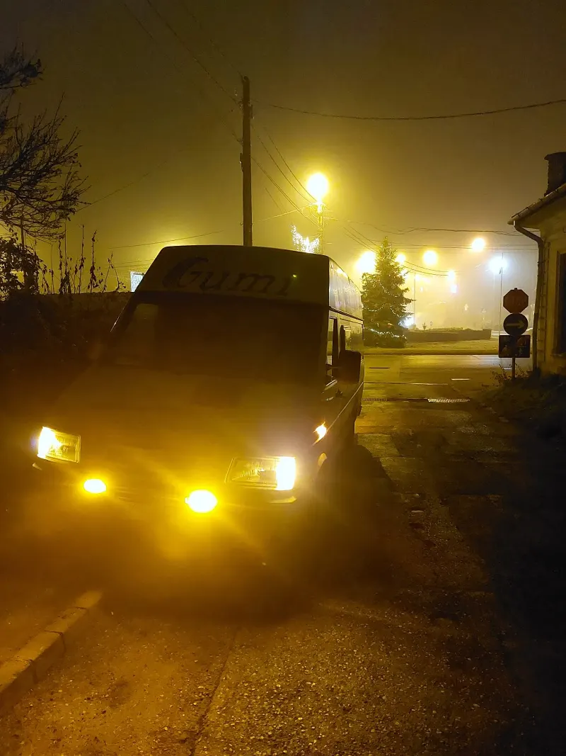 A szervizbusz ködös éjszakában, felkapcsolt sárga lámpákkal.