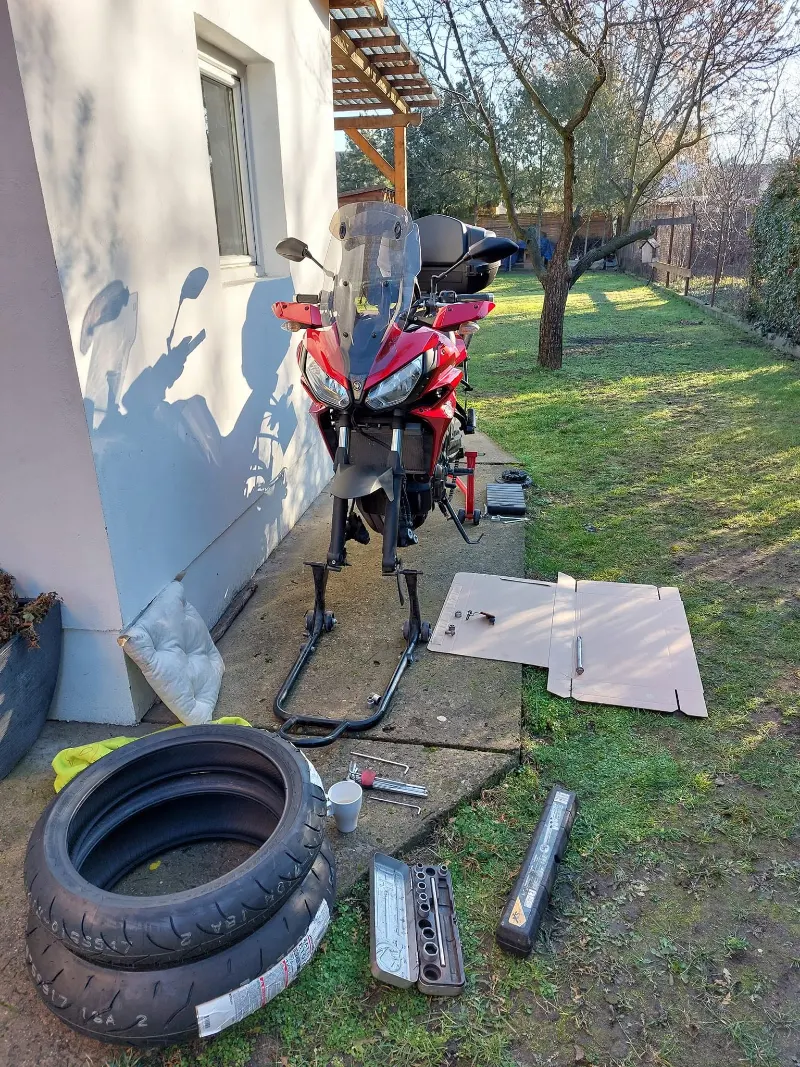 Motorkerékpár szerelőállványon egy kertben, mellette az új gumik.