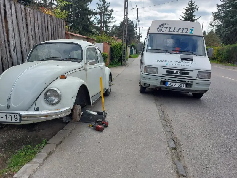 Fehér veterán VW Bogár emelőn az utcán, mögötte a szervizbusz.